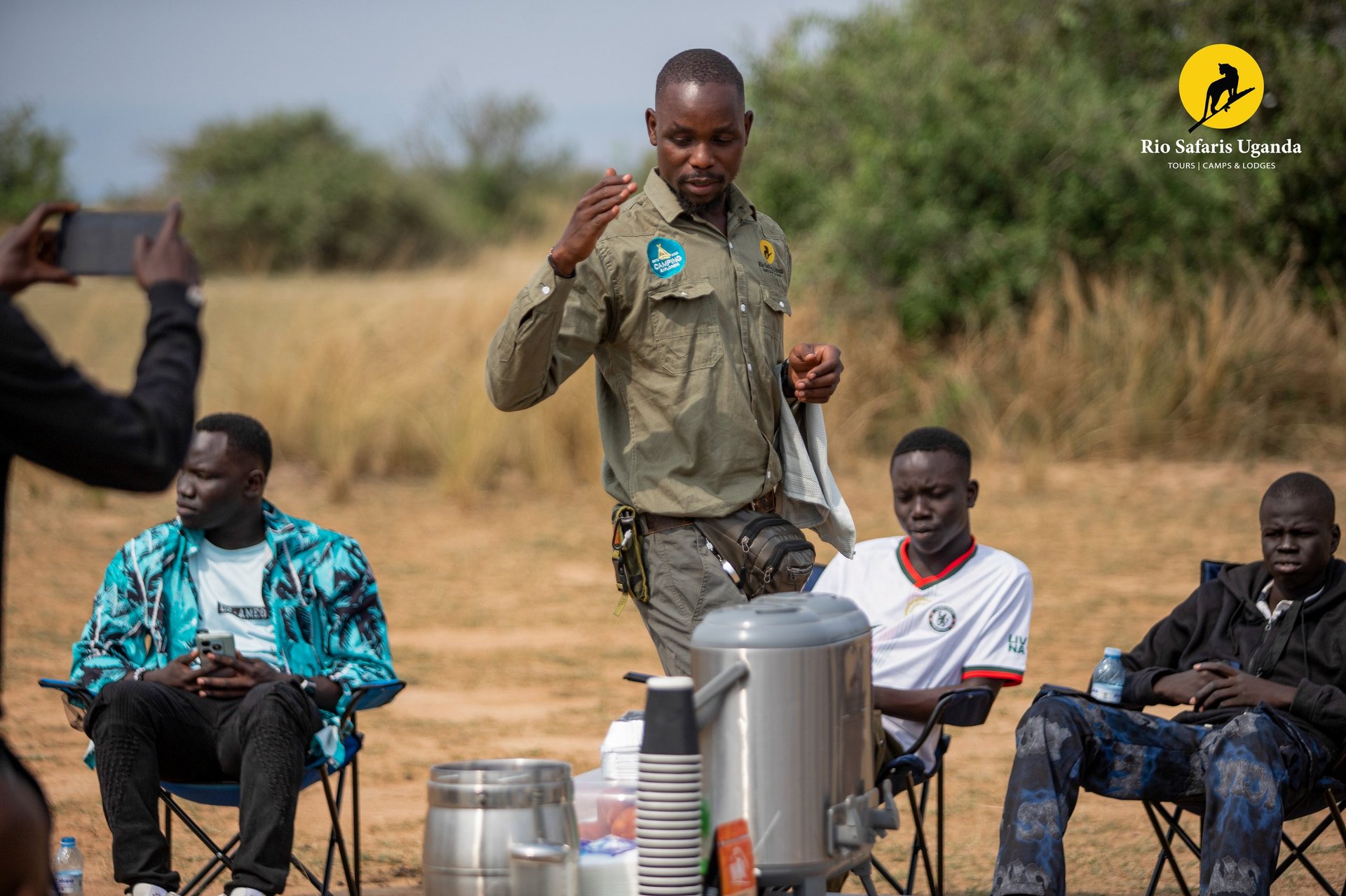 Tour guide demonstrating cooking equipment to seated visitors outdoors in a savanna setting
