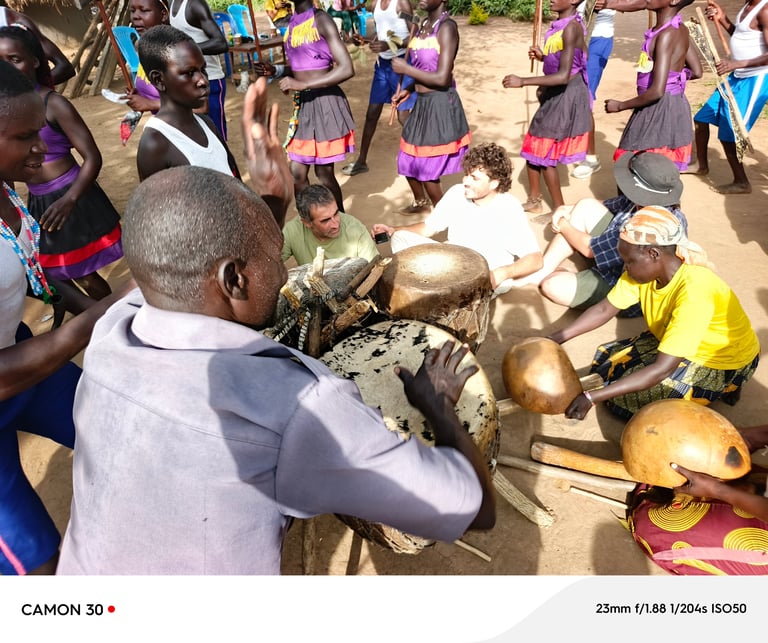 Community members gather around large drums in an outdoor celebration, with people in colorful purple and pink traditional attire playing and dancing