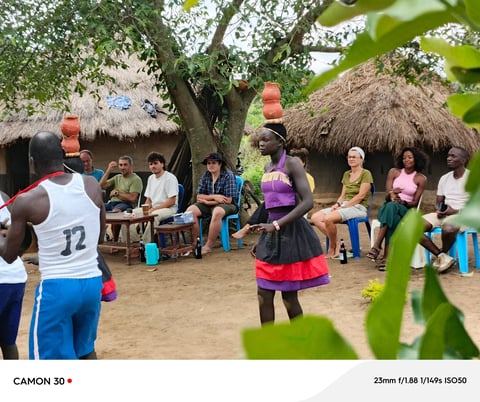 Group of people gathered in a village compound with thatched-roof structures, watching a soccer player in action during a community match