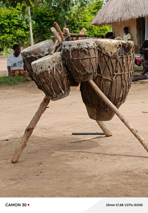 Traditional wooden drums suspended on wooden legs in an African village setting with thatched-roof structures in background