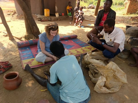 Adults and children sitting on a colorful mat on the ground in a rural village setting, engaged in an educational or community activity