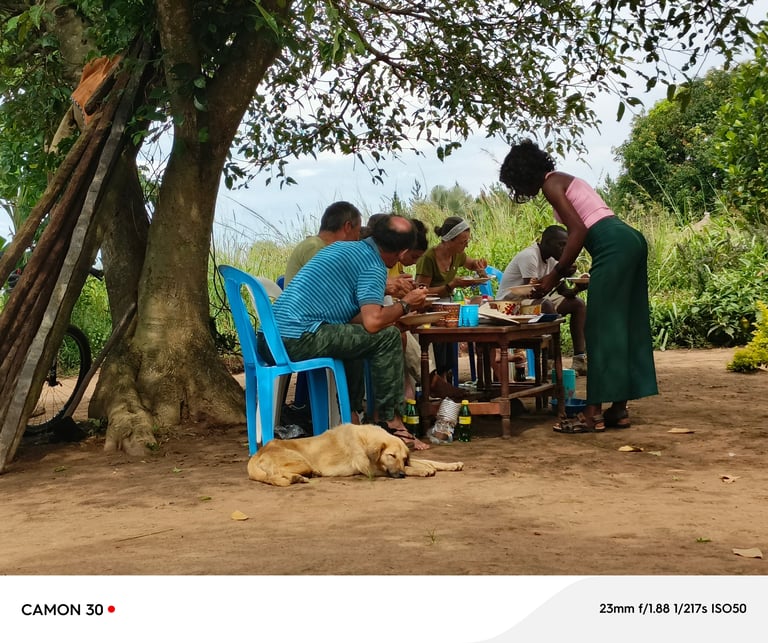 Family gathered under a large tree sharing a meal outdoors in a rural setting with green vegetation