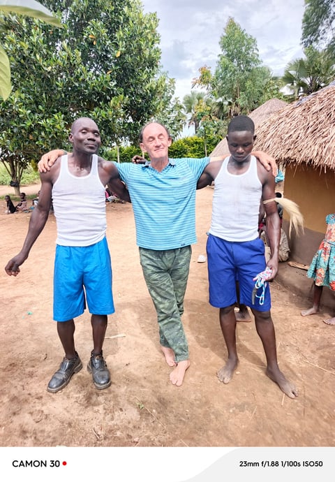 Three men standing together with arms around each other in a village setting with traditional huts and green trees in the background