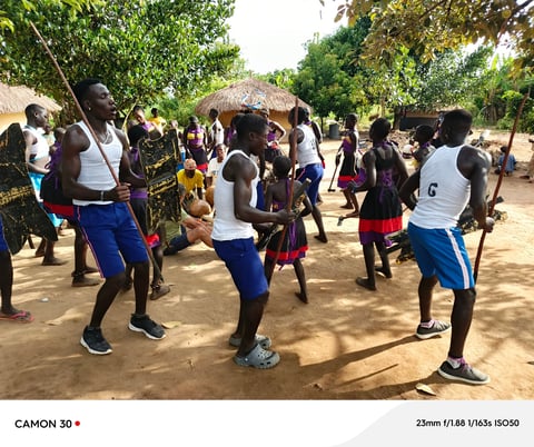 Group of children dancing and playing together in a rural African village courtyard with thatched-roof structures and trees