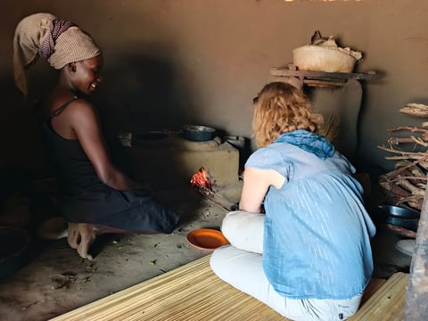 Two people cooking over a small fire in a basic kitchen, with pots and cooking utensils visible