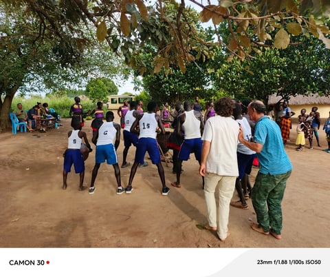 Youth playing volleyball on dirt court in rural village setting with spectators watching under trees