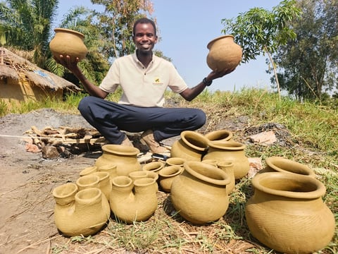 Man sitting among handmade clay pottery vessels, holding two large pots above his head and smiling at the camera outdoors