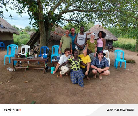 Group of people posing together in a rural village setting with thatched-roof huts and blue plastic chairs in the background