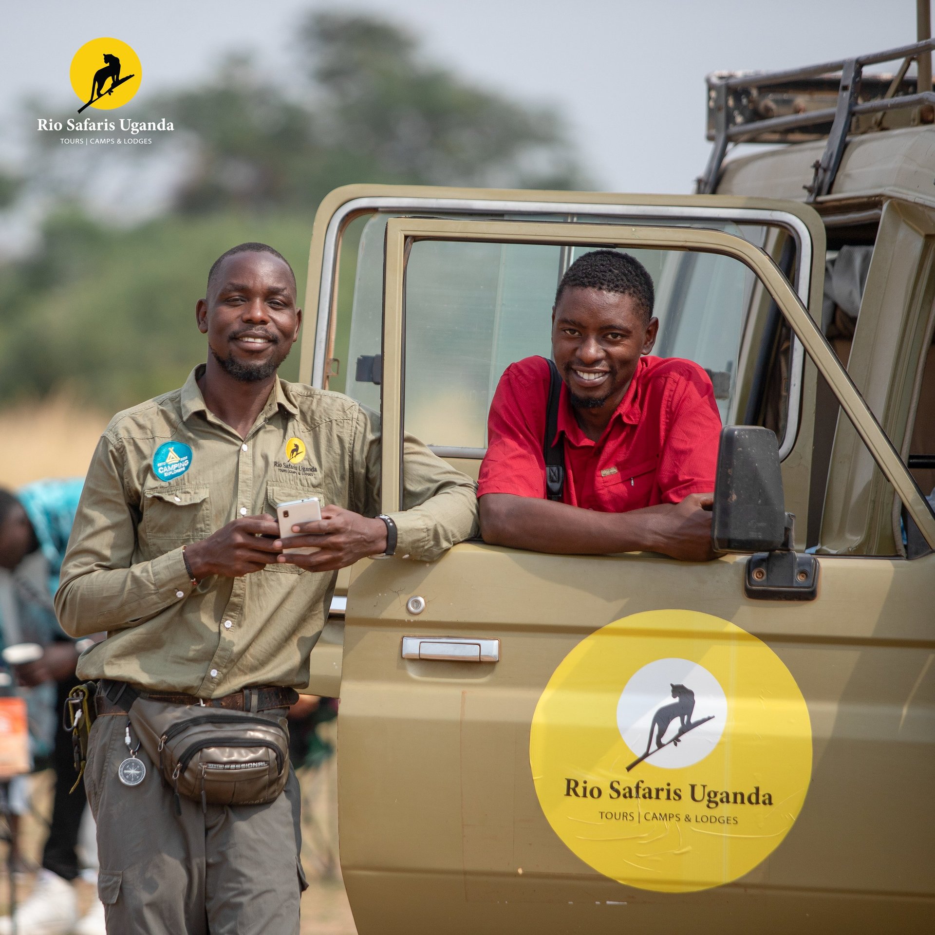 Two smiling staff members stand beside a tan Rio Safaris Uganda vehicle with yellow branding and safari logos