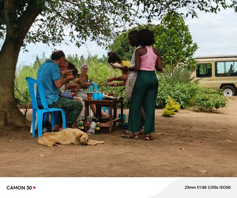 Family sitting outdoors under a tree sharing a meal, with a dog resting nearby and a vehicle in the background