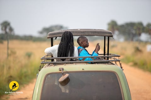 Two people on the roof of a safari vehicle on a dirt road in an African savanna landscape