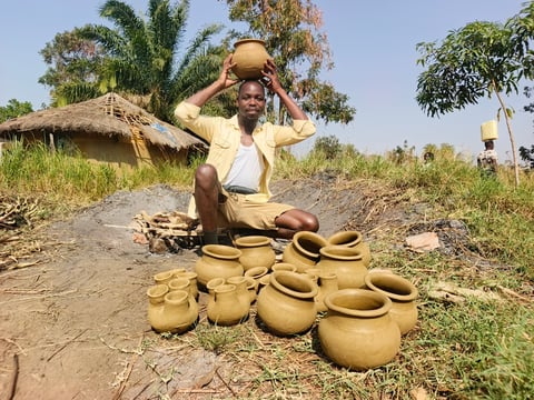 Man in yellow shirt displaying handmade clay pots and vessels in rural village setting with thatched hut