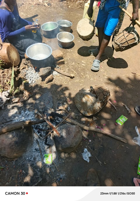 Outdoor cooking scene with metal pots arranged on the ground near a campfire, people standing nearby on dirt ground