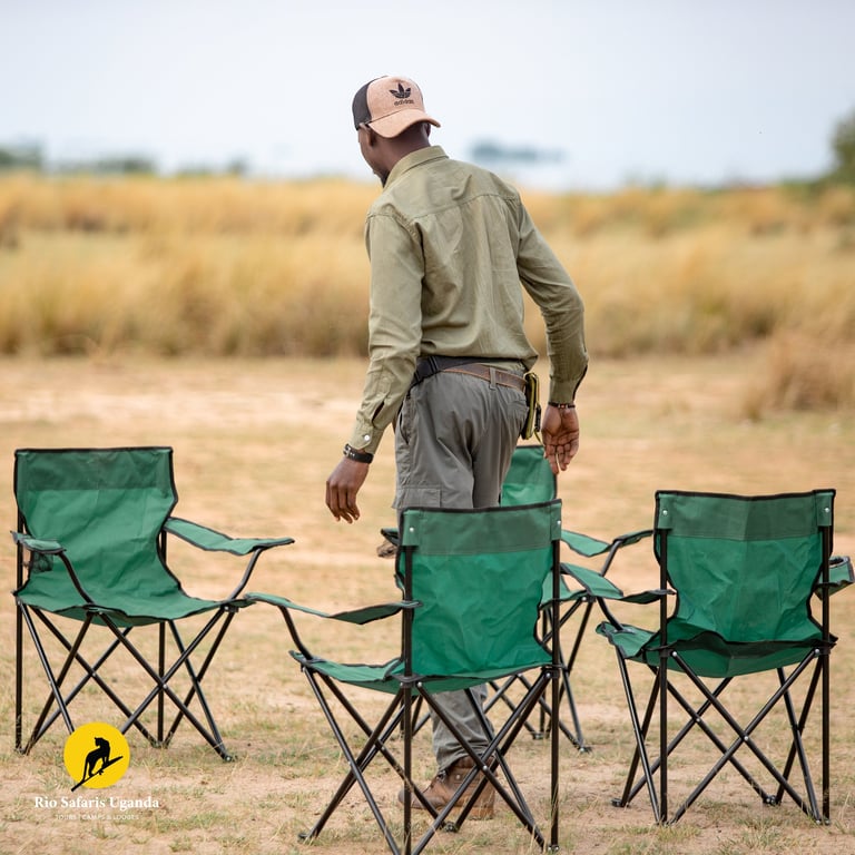 Man standing in desert landscape surrounded by three green camping chairs, wearing tan jacket and cap