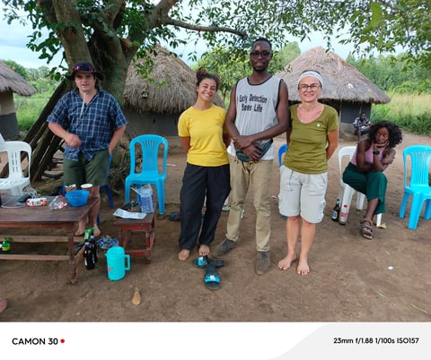 Group of six people standing outdoors in a village setting with thatched-roof huts and blue plastic chairs visible in the background