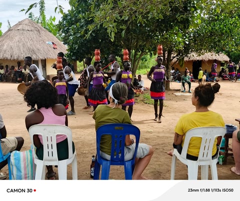 Community gathering in African village with children and adults in colorful clothing standing in sandy plaza near thatched-roof structures