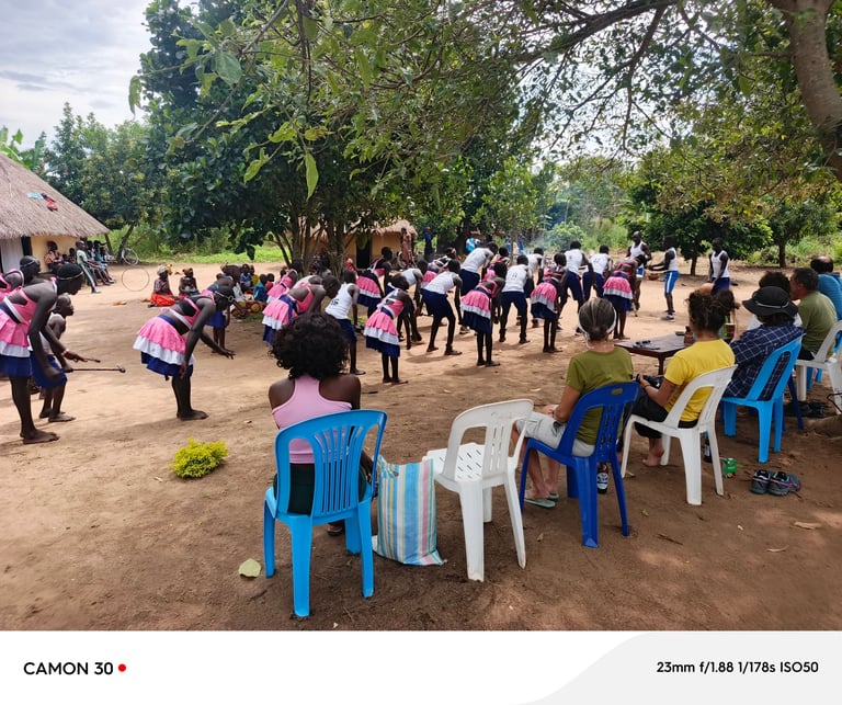 Community gathering in rural village with children wearing pink uniforms and adults seated on blue plastic chairs under trees