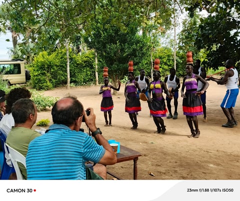 Group of children in purple uniforms performing a dance or cultural activity outdoors while spectators watch and photograph the event