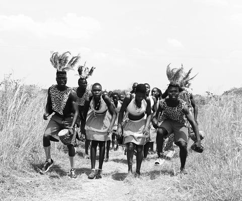 Group of people in traditional African attire and headdresses walking through grassland in black and white photograph