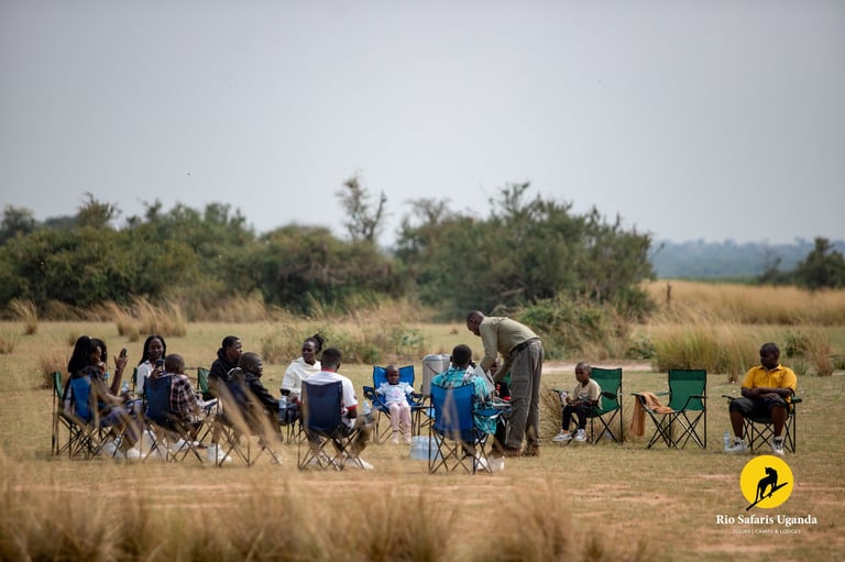 Group of people sitting in camping chairs in a grassland with trees in the background during what appears to be a safari or outdoor gathering