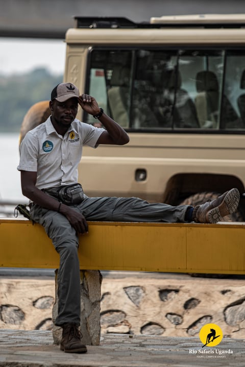 Safari guide in white uniform sits on yellow barrier with safari vehicle in background, adjusting his cap