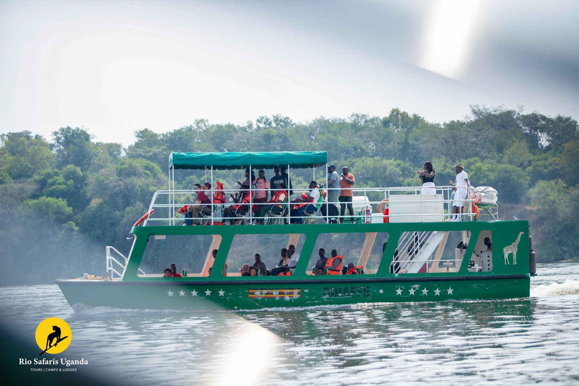 A green and blue safari boat filled with tourists on the water with forested shoreline in the background