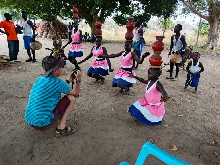 Group of African children playing in a circle outdoors under trees, wearing colorful clothing and protective gear on dusty ground