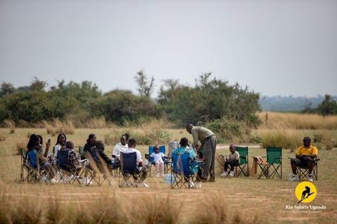 Group of tourists in camping chairs on sandy ground with trees in background during wildlife safari