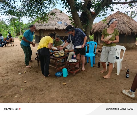 Group of visitors and locals gathered around a table in a rural village with traditional thatched-roof huts