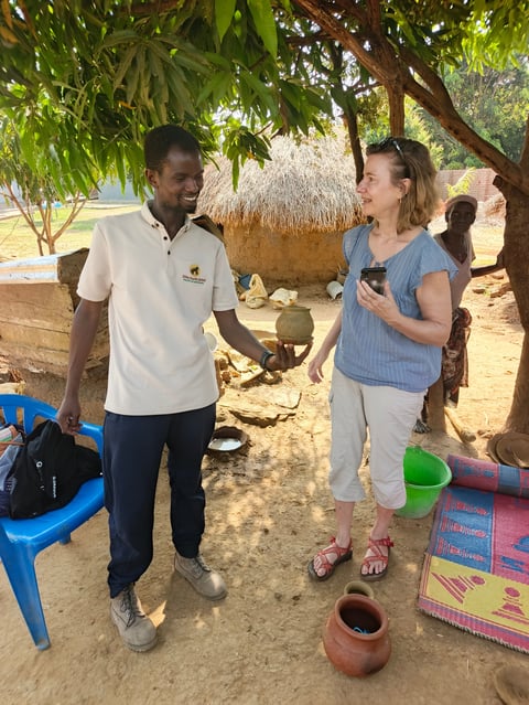 Man in white polo shirt and woman in blue shirt conversing under a large tree in a sandy village setting with traditional huts