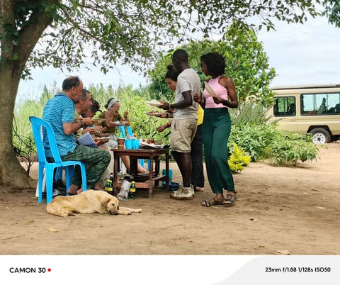 Group of people sharing a meal outdoors under a large tree with a safari vehicle parked in the background
