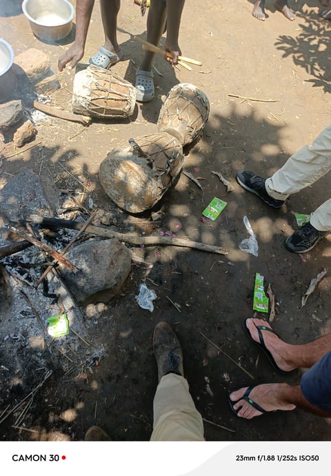 Overhead view of food being prepared in pots over a fire pit on sandy ground with people standing around