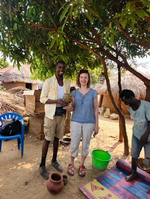 Woman standing with two men in a rural African village setting under a large tree, with thatched-roof huts and pottery visible