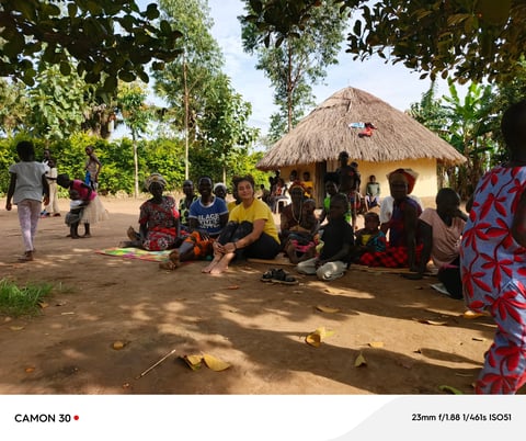 Group of people gathered outside a thatched-roof hut in an African village, sitting on the ground in a community gathering