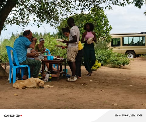 Group of people gathered under a tree with a brown vehicle in background, dog resting on ground, rural setting
