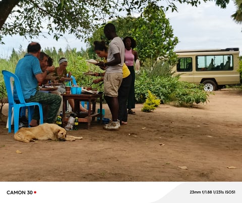 A group of people gathered outdoors around a table and cooking area near a van in a rural setting with green vegetation