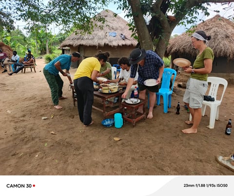 Community gathering around a table sharing a meal outdoors near traditional thatched-roof huts in a village setting
