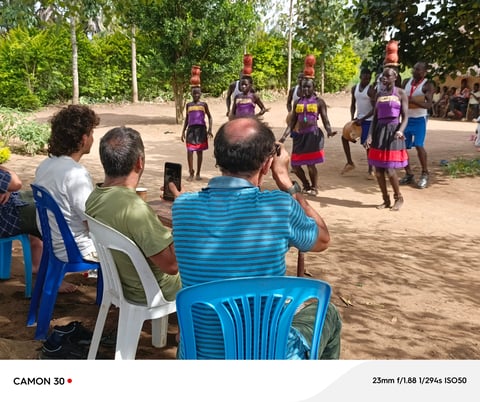 Spectators seated on blue and white plastic chairs watch a group of young dancers performing in traditional purple and red clothing in an outdoor courtyard